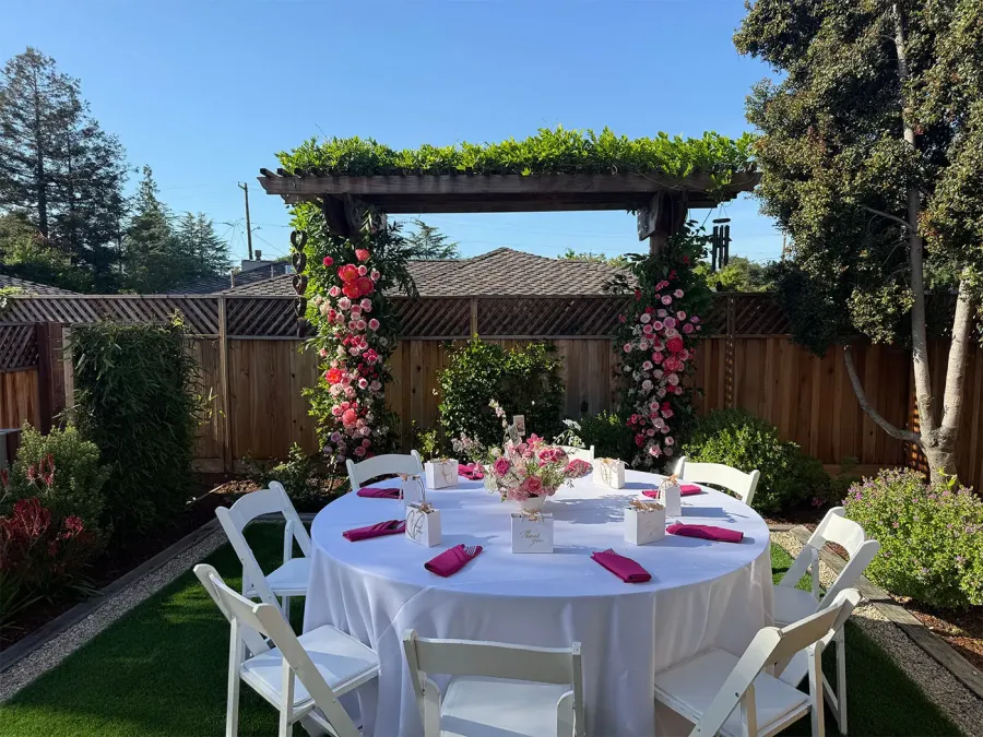 Rose arch in blush and deep red framing a round garden table finished in white linen with fuchsia napkins — floral install styling from our Livermore catering and florals team